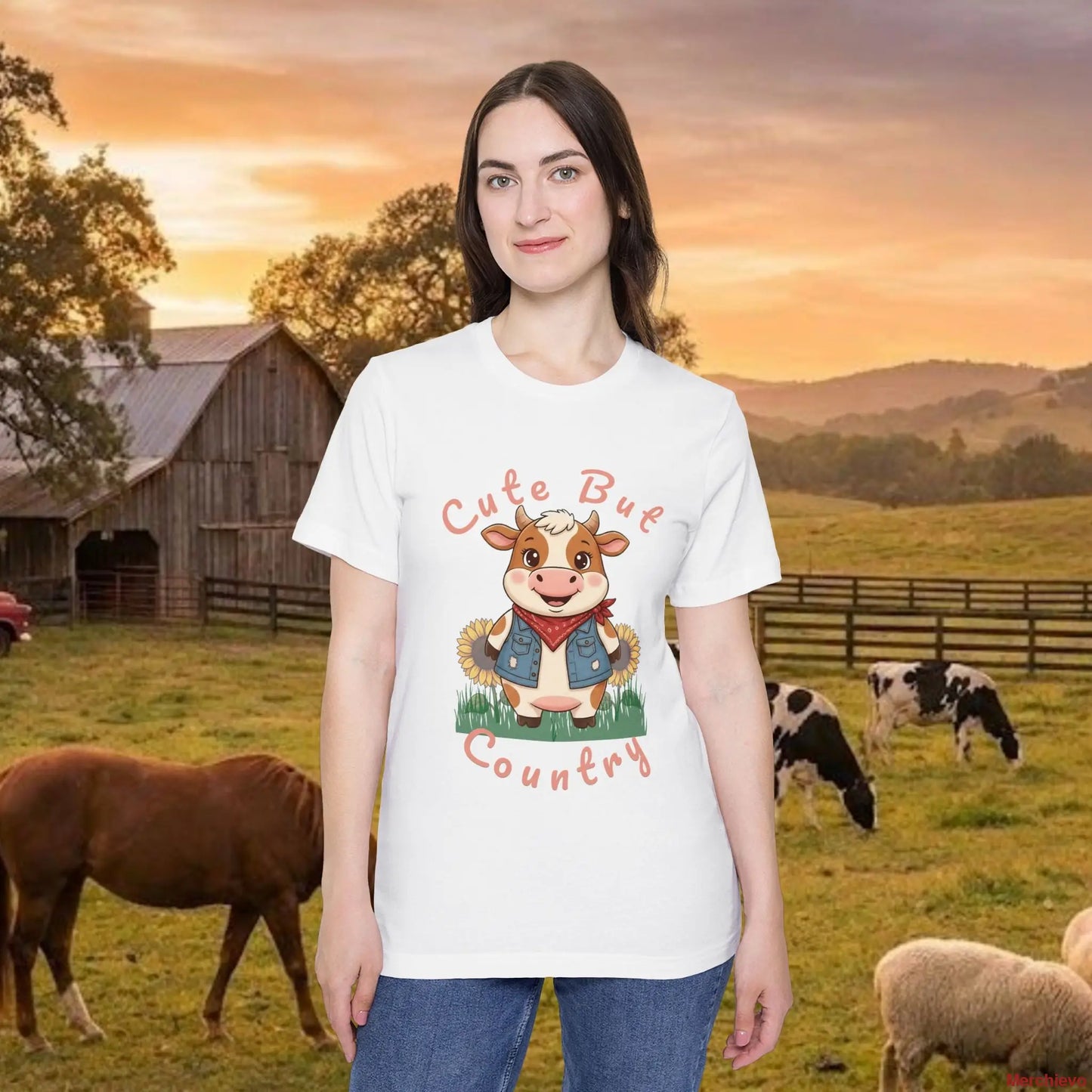A woman wearing a white t-shirt with a cartoon graphic of a smiling brown cow. The cow is wearing a red bandana and a denim vest, surrounded by sunflowers and the text "Cute but Country." She is standing in a farm field with a barn, cows, and a horse in the background during a golden hour sunset.